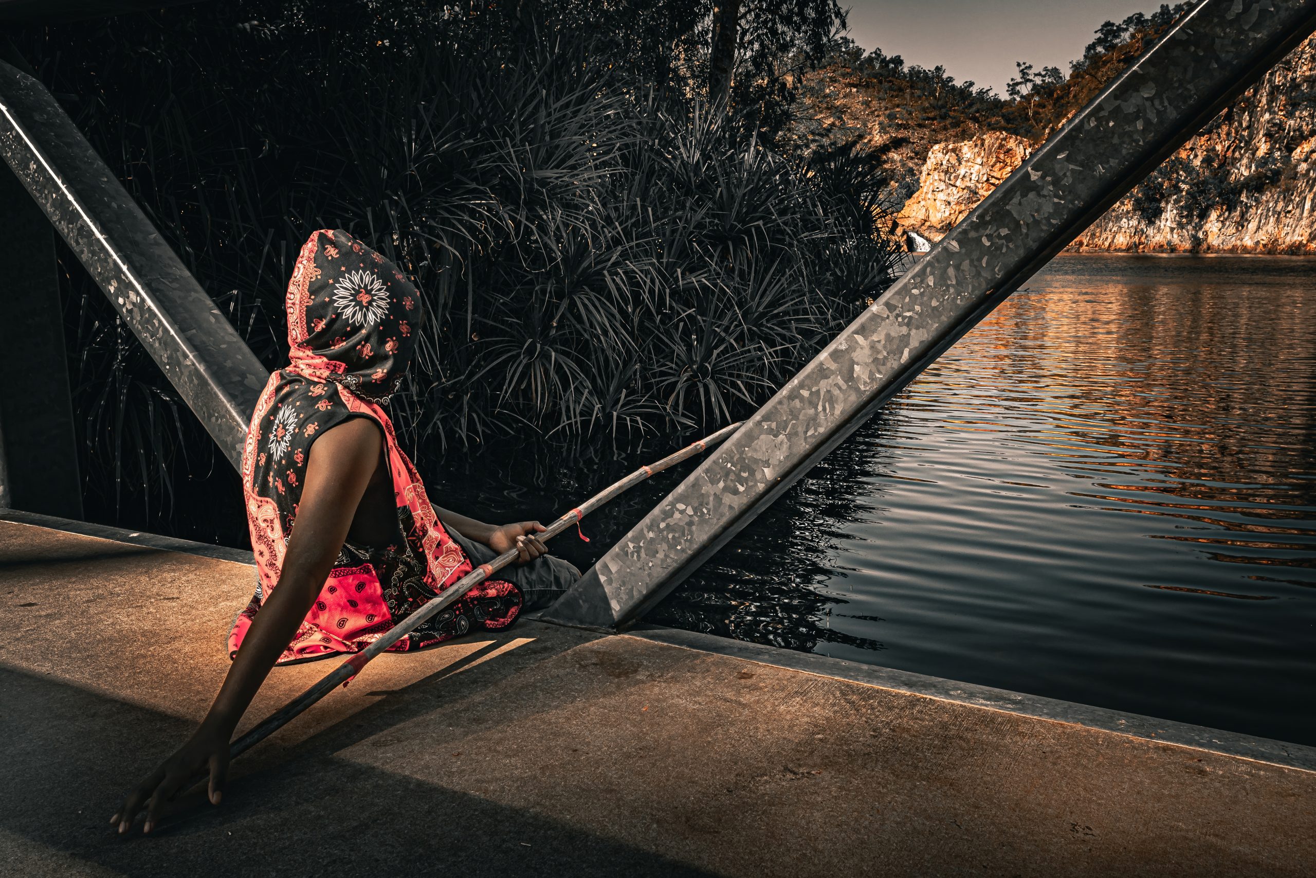 A person sitting by the water holding a stick, dressed in a colorful hood and scarf, with lush greenery and rocky cliffs in the background.