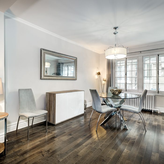 A modern dining room with a glass table, grey chairs, hardwood floors, and large windows allowing natural light to fill the space.