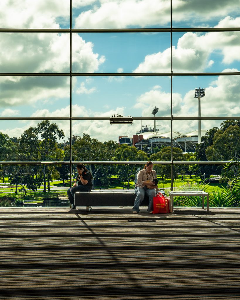 Two people seated on a bench in front of a large glass window, with a view of the outdoors and a sports stadium in the background.