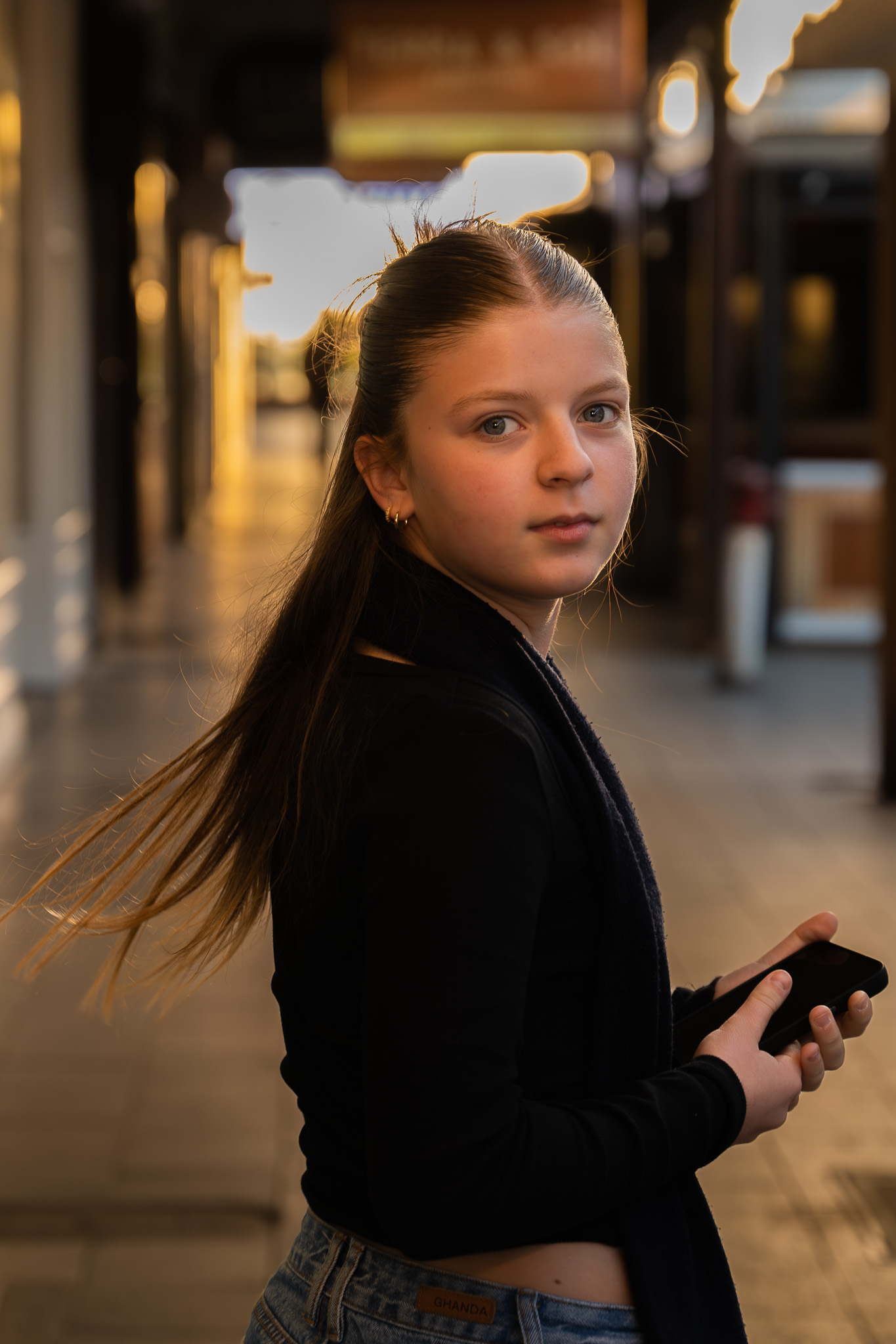 Portrait of a young girl holding a phone, captured outdoors with soft golden light in the background.