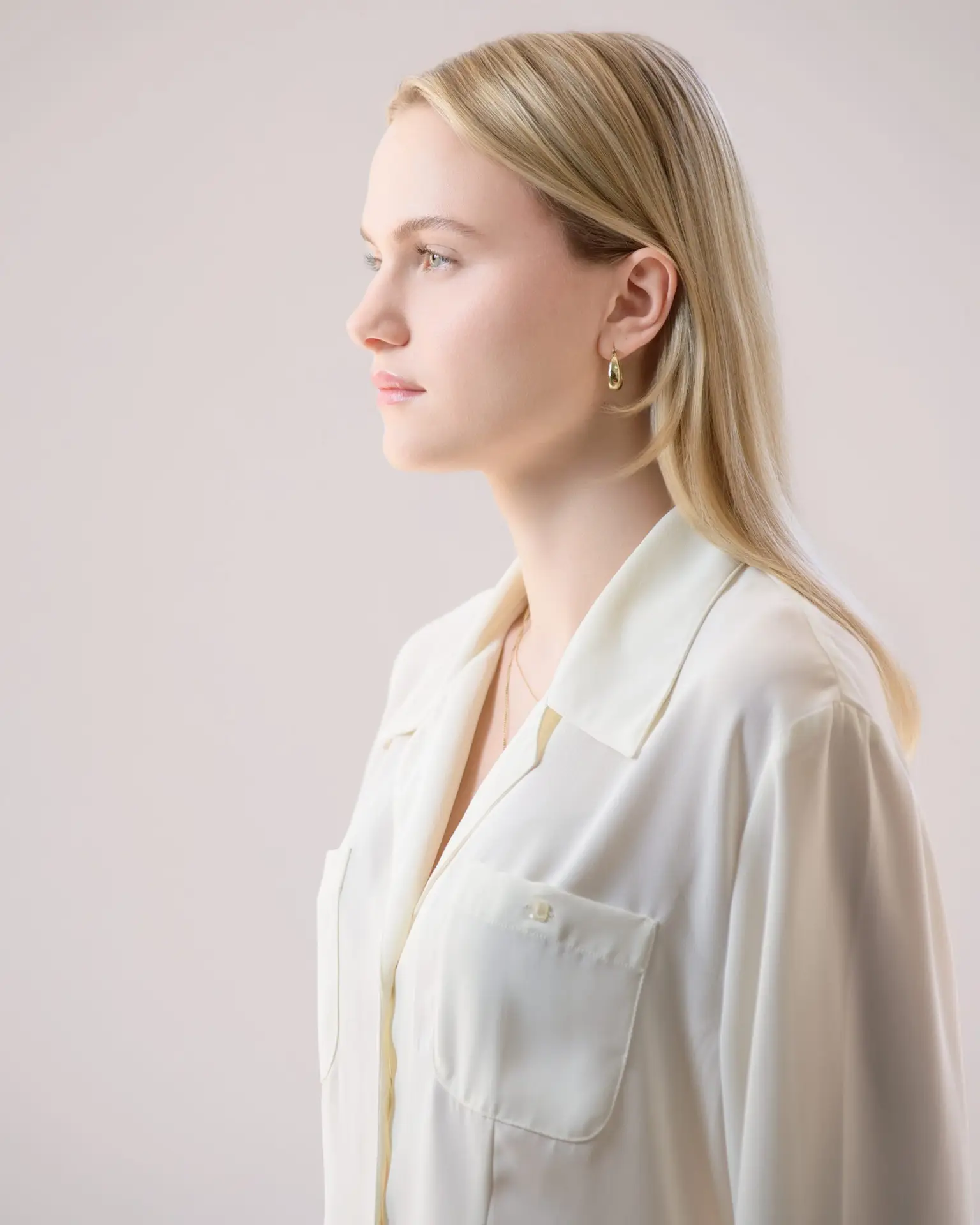 Profile portrait of a young woman with blonde hair, wearing a cream blouse, looking to the left, set against a soft, light background.