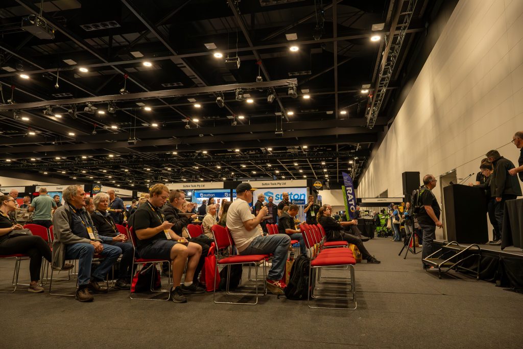 Audience seated at a corporate event with a presentation stage and exhibition booths in the background.