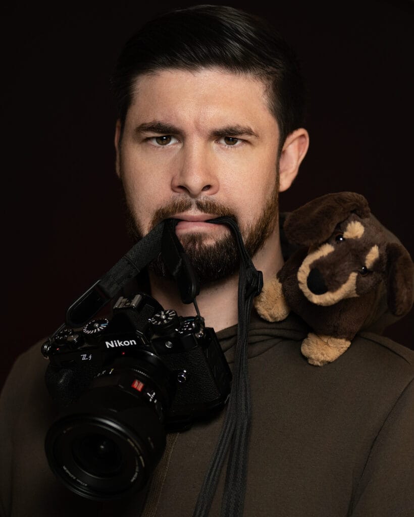 Corey, Adelaide photographer and videographer, holding a Nikon camera with a toy dachshund on his shoulder — representing the personality behind Shameless Visuals.