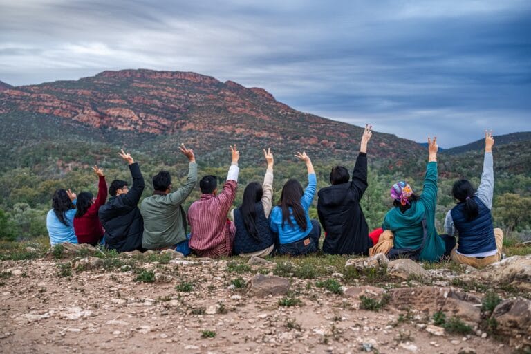 Australia Awards scholars seated on a ridge lookout at Wilpena Pound with hands raised toward the Flinders Ranges