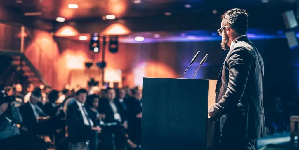 A speaker presenting at a corporate event, with an audience seated in the background.